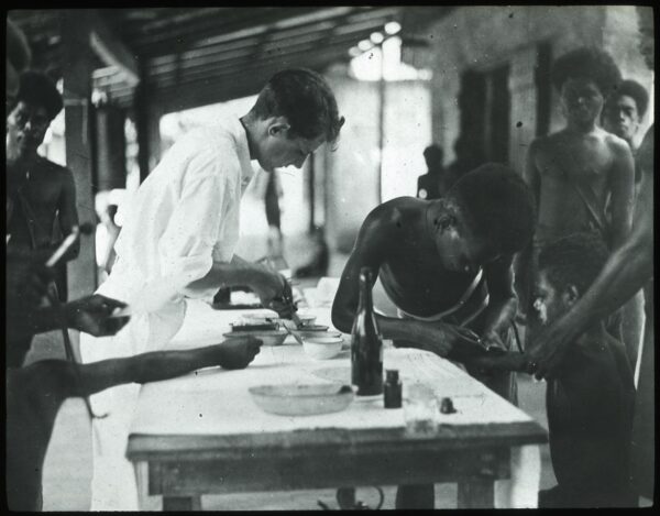 A black and white photograph. Under a covered awning, a white doctor in a white shirt and white pants bends over a table. He holds a needle in his hand and appears to fill the needle with the contents of a bowl. A Solomon Islander on the other side of the table injects a young boy, and other Solomon Islanders surrond the table and watch.