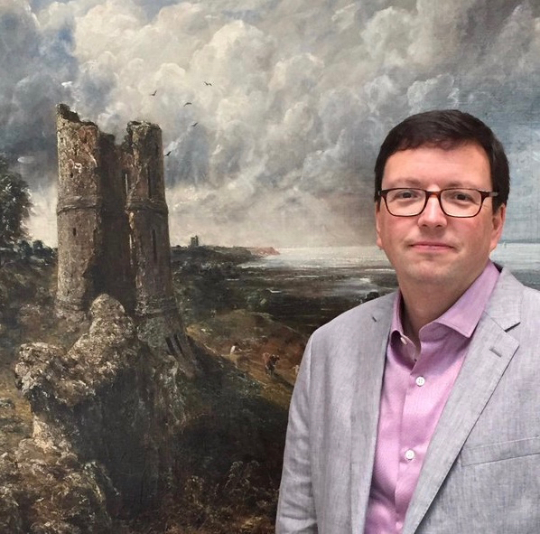 A middle-aged man with dark hair and glasses stands in front of a painting of a turbulent landscape.