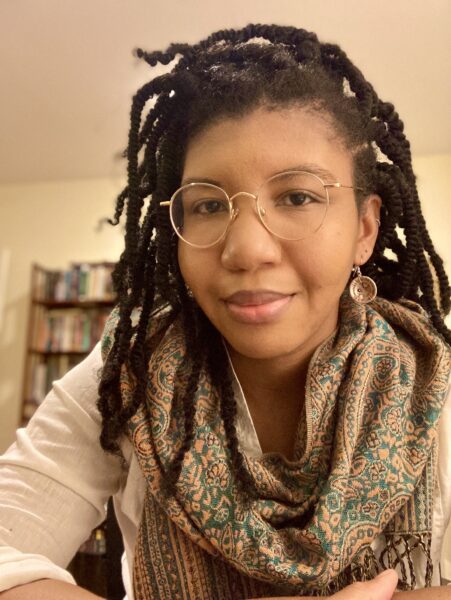 A headshot of Dr. Elise A. Mitchell wearing a white blouse and gold and teal scarf. She is sitting in front of a bookcase.