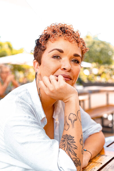 A Black mixed race woman with short peach coloured hair and a tattooed forearm rests her elbows on a table and has her hand on her chin. She is smiling. 