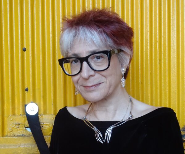 Image is of a short white woman in her sixties, with red-and-white striped hair and angular black glasses. She is smiling and wears a black sweater with a wide neck, a silver necklace in the shape of skeleton hands, and silver earrings. She stands outside in front of a bright-yellow elevator door.